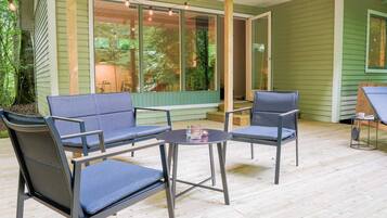 Un patio exterior con dos sillas y una mesa, un área cubierta con luces colgantes, y vista a una casa verde con ventanas grandes.