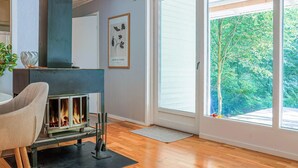 A modern living room with a fireplace, a dining area with a chair, and a large sliding glass door leading to a green outdoor area. - 4 Person Holiday Home in Horby (Horby)