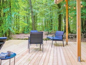 An outdoor deck with wooden flooring, surrounded by a forest. There are two chairs and a table with a book and a mug. - 4 Person Holiday Home in Horby (Horby)