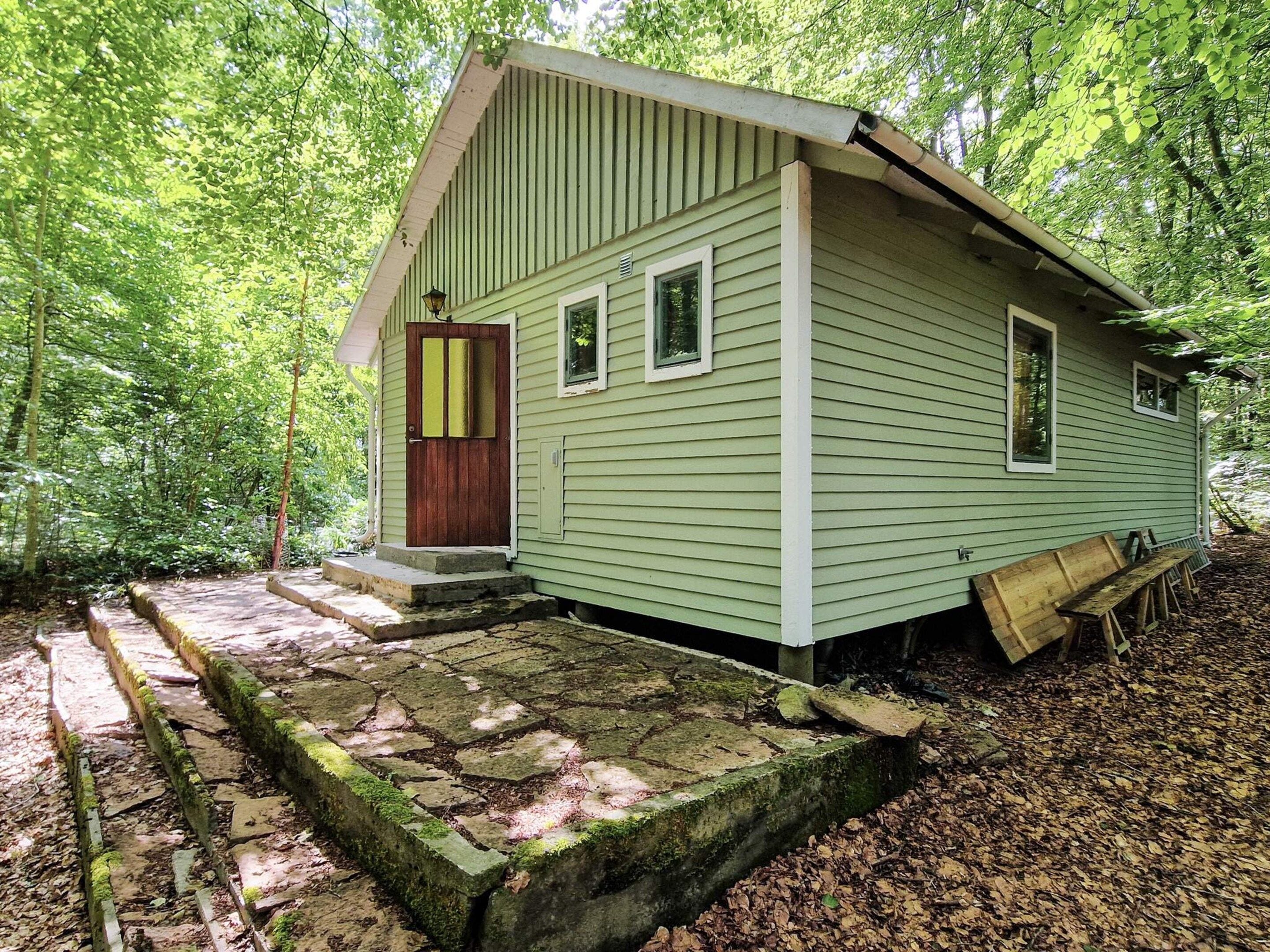 Une petite cabane à un seul étage, dotée d’une porte d’entrée et de fenêtres, entourée d’un sentier pavé de pierres et d’une forêt.