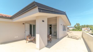 En moderne bygning med fladt tag, en overdækket patio med to liggestole og et bord, samt en skydedør til et indre rum.
