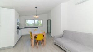 Et moderne køkken med hvide skabe, et træspisebord med gule stole, en grå sofa og et stort vindue, der giver naturligt lys.