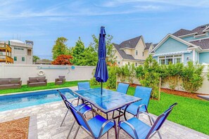 Ein Garten mit Schwimmbecken, Terrassentisch und Stühlen sowie einem blauen Sonnenschirm.