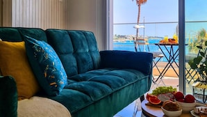 Een kamer met een groene bank, een kleine tafel met fruit en drankjes, en uitzicht op zee en palmbomen.