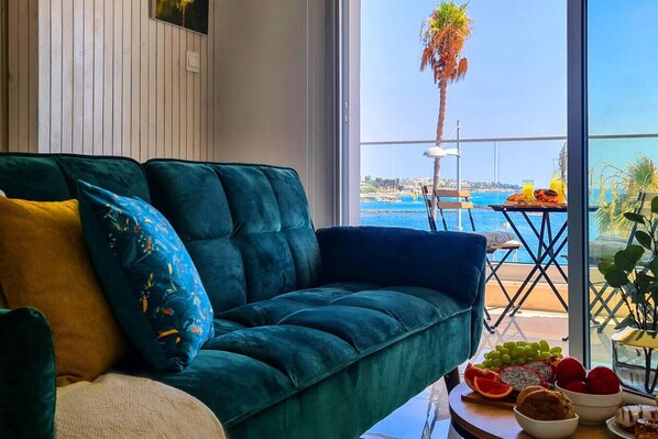 Quarto com sofá verde, uma pequena mesa com frutas e bebidas, e vista para o mar e palmeiras.