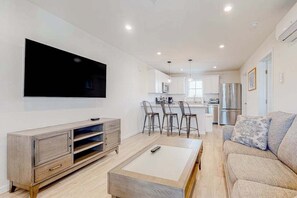 A modern living room with a flat-screen TV, a wooden coffee table, a sofa, and a kitchen area in the background. - Rehoboth Beach House condo with dazzling interior, pool access - nearby golf (Rehoboth Beach)