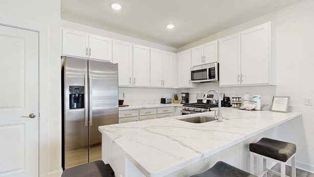 A modern kitchen with white cabinets, a stainless steel refrigerator, a microwave, and a large island with bar stools. - Bayfront Condo | View, Near Lewes Beaches (Lewes)