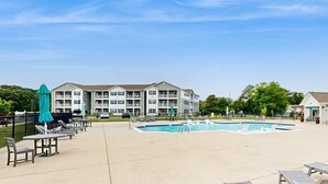 A pool area with lounge chairs and umbrellas, a building with multiple floors, and a clear sky. - Bayfront Condo | View, Near Lewes Beaches (Lewes)