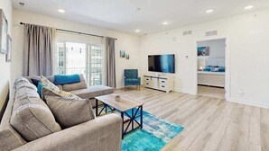 A modern living room with a sofa, a coffee table, a TV, and a kitchen area visible through an open door. - Bayfront Condo | View, Near Lewes Beaches (Lewes)