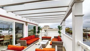 A covered outdoor seating area with wicker furniture and orange cushions, a glass door leading inside, and a view of buildings in the background. - Town of Rehoboth Beach apartment w/balcony, walk to beach (Rehoboth Beach)