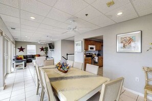 Un coin repas avec une table, des chaises et un ventilateur de plafond. On aperçoit une cuisine en arrière-plan, avec des placards et un four à micro-ondes.