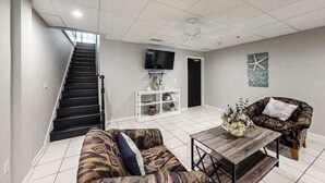 A living room with a staircase, a flat-screen TV, a white console table, a patterned sofa, a wooden coffee table, and a decorative wall piece. - Town of Rehoboth Beach apartment w/balcony, walk to beach (Rehoboth Beach)