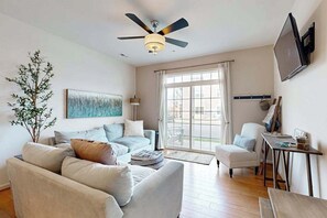 A living room with a ceiling fan, a sofa, a chair, a table, and a TV mounted on the wall. - Grande at Canal Pointe pet friendly condo w/shared pool, tennis courts & gym (Rehoboth Beach)