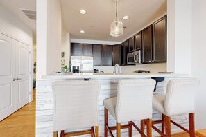 A modern kitchen with dark cabinets, a white island, and three bar stools. - Grande at Canal Pointe pet friendly condo w/shared pool, tennis courts & gym (Rehoboth Beach)