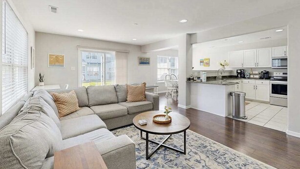 A spacious living room with a gray sectional sofa, a round coffee table, and a kitchen area with white cabinets and appliances. - Eagles Landing townhome with pool and tennis access, patio & grill (Rehoboth Beach)