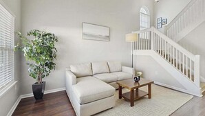 A living room with a beige sofa, a wooden coffee table, a potted plant, a lamp, and a staircase. - Eagles Landing townhome with pool and tennis access, patio & grill (Rehoboth Beach)