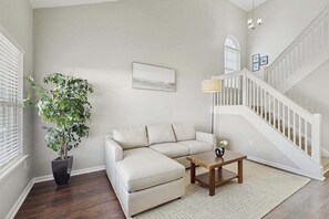 Ein Wohnzimmer mit einem beigen Sofa, einem Holztisch, einer Zimmerpflanze, einer Lampe und einer Treppe.
