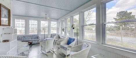 Une véranda avec des meubles en osier, de grandes fenêtres et un ventilateur de plafond.