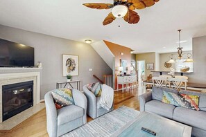 A modern living room with a fireplace, grey sofas, a ceiling fan, and a dining area in the background. - Rehoboth Crossing townhome - pool & gym - dog-friendly, fireplace & private W/D (Rehoboth Beach)