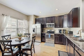 A modern kitchen with dark cabinets, stainless steel appliances, a dining area with wooden chairs, and a window with sheer curtains. - Rehoboth Crossing townhome - pool & gym - dog-friendly, fireplace & private W/D (Rehoboth Beach)