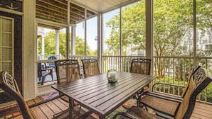 A covered patio with a wooden table and wicker chairs, large windows, and a view of trees and buildings. - Bear Trap Dunes 1st floor condo w/ tennis court, lakefront, and golf on-site (Ocean View)