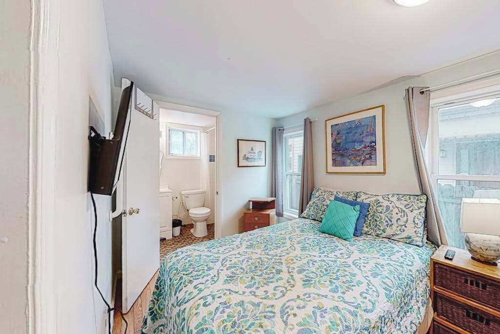 A bedroom with a bed, a bedside table, a window with curtains, a painting on the wall, and a bathroom visible through an open door.