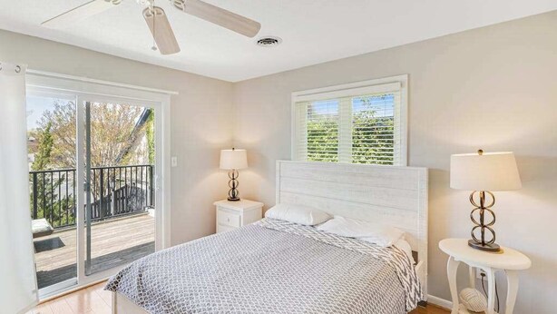 Une chambre à coucher comprenant un lit, un ventilateur de plafond, une fenêtre avec des stores, une table de chevet avec une lampe et une porte-fenêtre donnant sur un balcon.
