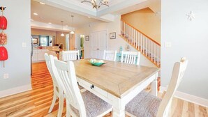 A dining area with a wooden table and white chairs, a staircase, and a kitchen in the background. - Lovely, dog-friendly townhouse near the beach with a pool & washer/dryer (Rehoboth Beach)