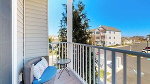 A balcony with a wicker chair and a small table, overlooking a street with houses and a clear sky. - Grande at Canal Pointe condo with pools, tennis, gym, & private balcony (Rehoboth Beach)
