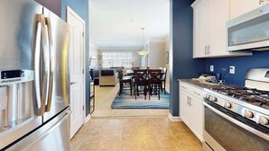 A modern kitchen with stainless steel appliances, a dining area with a blue rug, and a view into the living room. - Grande at Canal Pointe condo with pools, tennis, gym, & private balcony (Rehoboth Beach)