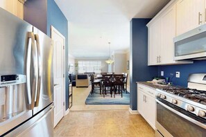 A modern kitchen with stainless steel appliances, a dining area with a blue rug, and a view into the living room. - Grande at Canal Pointe condo with pools, tennis, gym, & private balcony (Rehoboth Beach)