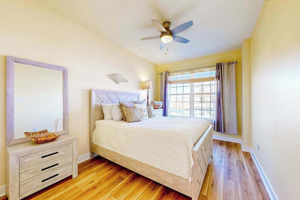 A bedroom with a bed, a ceiling fan, a window with curtains, and a bedside table with a lamp.