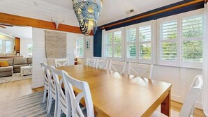 Ein Essbereich mit Holztisch und weißen Stühlen, einer Zierleuchter und großen Fenstern mit Jalousien.