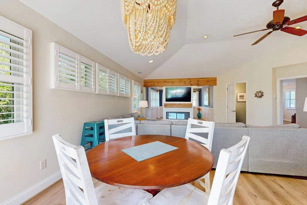 Un vaste espace de vie comprenant une table à manger en bois, des chaises blanches, un ventilateur de plafond et un grand téléviseur.