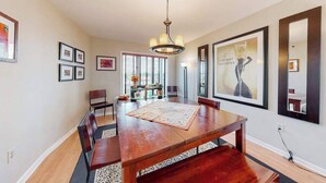 A dining room with a wooden table, chairs, a chandelier, framed artwork, and a window with blinds. - Town of Rehoboth Beach, Wilmington Ave #317 | Condo in Rehoboth Beach (Rehoboth Beach)