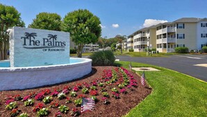 A sign for 'The Palms of Rehoboth' with a landscaped area featuring flowers and an American flag. - The Palms condo near the beach with on-site pool & enclosed patio (Rehoboth Beach)