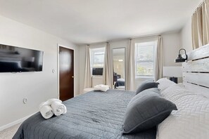 Ein Schlafzimmer mit Bett, einem an der Wand befestigten Fernseher und einem Fenster mit Vorhängen.