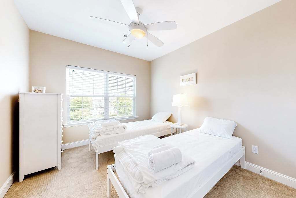 A hotel room with two beds, a ceiling fan, a window with blinds, and a bedside lamp.