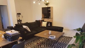 A modern living room with a sectional sofa, a coffee table, a printer, and a bookshelf. - L'écrin de l'abreuvoir – Cozy Ground-Floor Retreat in Marnay Near Besançon (Marnay)