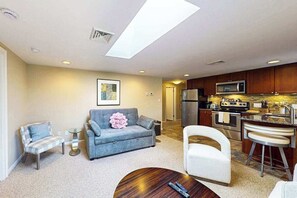 Una sala de estar con sofá, sillones, una mesa de centro y una cocina con electrodomésticos.