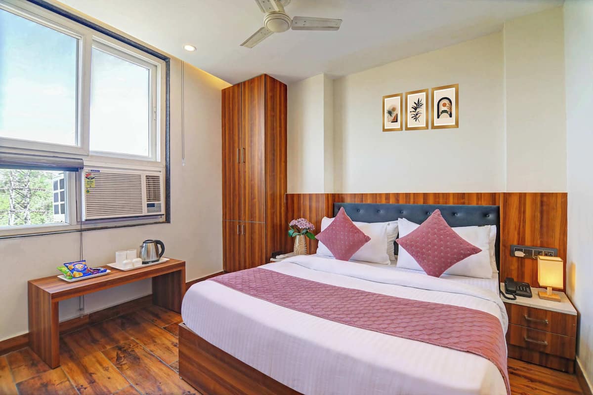 A hotel room with a large bed, wooden wardrobe, bedside table with a kettle, and a window with blinds.