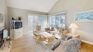 Un salón con sofá, sillón y televisor, una chimenea y ventanas grandes.