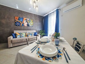 A dining area with a table set for two, a sofa with patterned cushions, a wall-mounted painting, and a potted plant. - Siciliandò by Interhome (Terrasini)