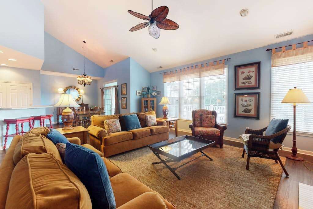 A spacious living room with a high ceiling, a ceiling fan, and a large window with curtains.