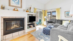 A living room with a fireplace, television, grey sofa, and a blue patterned rug. - Sea Colony Tennis second-floor condo with fireplace, gym, & tennis court (Bethany Beach)
