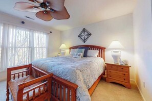 Ein Schlafzimmer mit Bett, Holzstuhl, Nachttisch, Lampe, Deckenventilator und einem Fenster mit Jalousien.