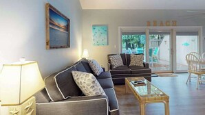 A living room with a sofa, a coffee table, and a lamp. There is a painting on the wall and a sign that reads 'BEACH'. - Sea Colony Tennis 1st-floor condo with balcony & gym (Bethany Beach)