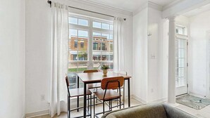 Een eetgedeelte met een tafel en stoelen, een raam met gordijnen en een uitzicht op gebouwen.