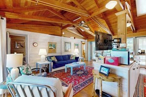 A cozy living room with a high ceiling, wooden beams, a blue sofa, a coffee table, a TV, and a kitchen area with cabinets and appliances. - Dewey Beach cottage 1 block from the beach, dog friendly (Dewey Beach)