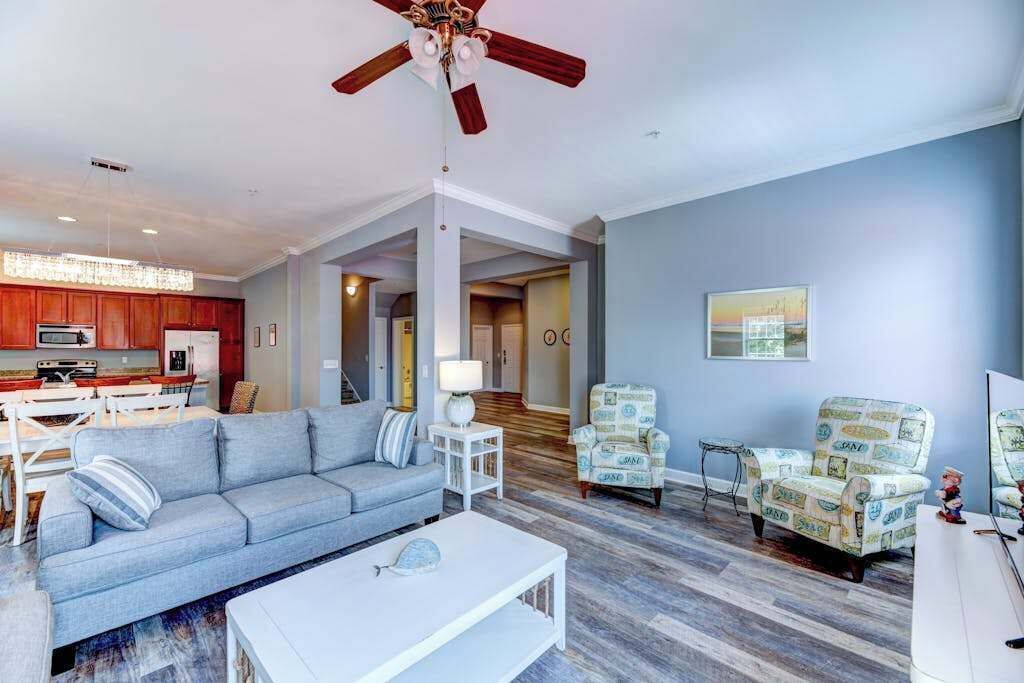 A spacious living area with a sofa, coffee table, patterned armchairs, a ceiling fan, and a kitchen in the background.